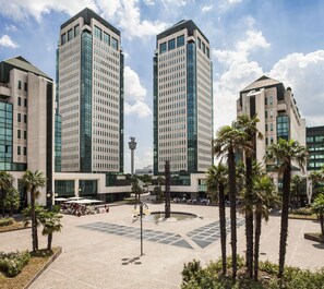 A modern urban plaza with tall buildings, palm trees, and outdoor seating areas. - Cosmo Residence (Vimercate)