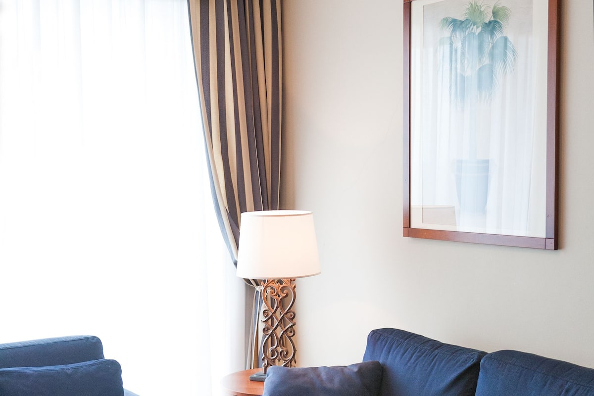 Un salon avec deux canapés bleu foncé, un plancher en bois, un ordinateur portable blanc posé sur le sol, une lampe sur une table d’appoint et un tableau encadré au mur.