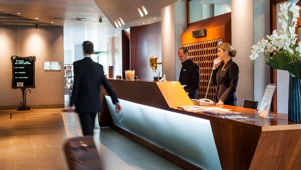 Een hotel receptie met een receptiebalie, een wandtelefoon en een bord met de lijst van kamer types.