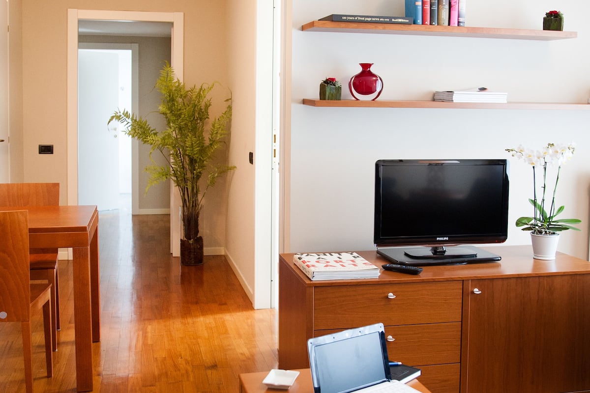 Un salon moderne avec un plancher en bois, un téléviseur posé sur un meuble, un ordinateur portable sur une table et une bibliothèque remplie de livres et d’objets décoratifs.
