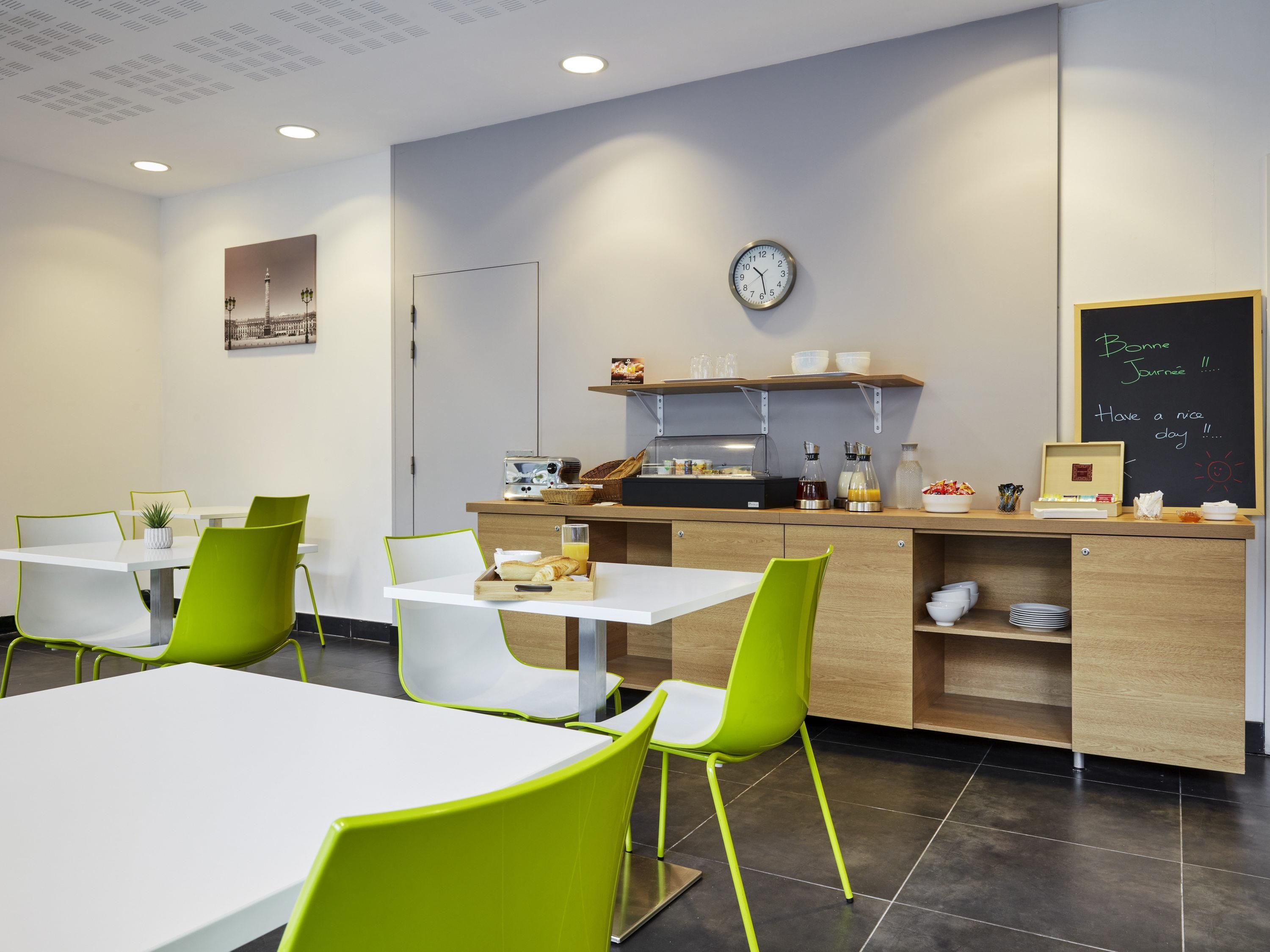 A modern cafe with white tables and green chairs, a clock, and a chalkboard with a message.