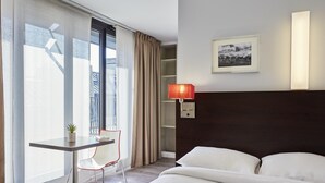 A hotel room with a bed, a desk with a chair, a window with curtains, a wall-mounted lamp, and a framed picture. - Aparthotel Adagio Access Paris Bastille (Paris)