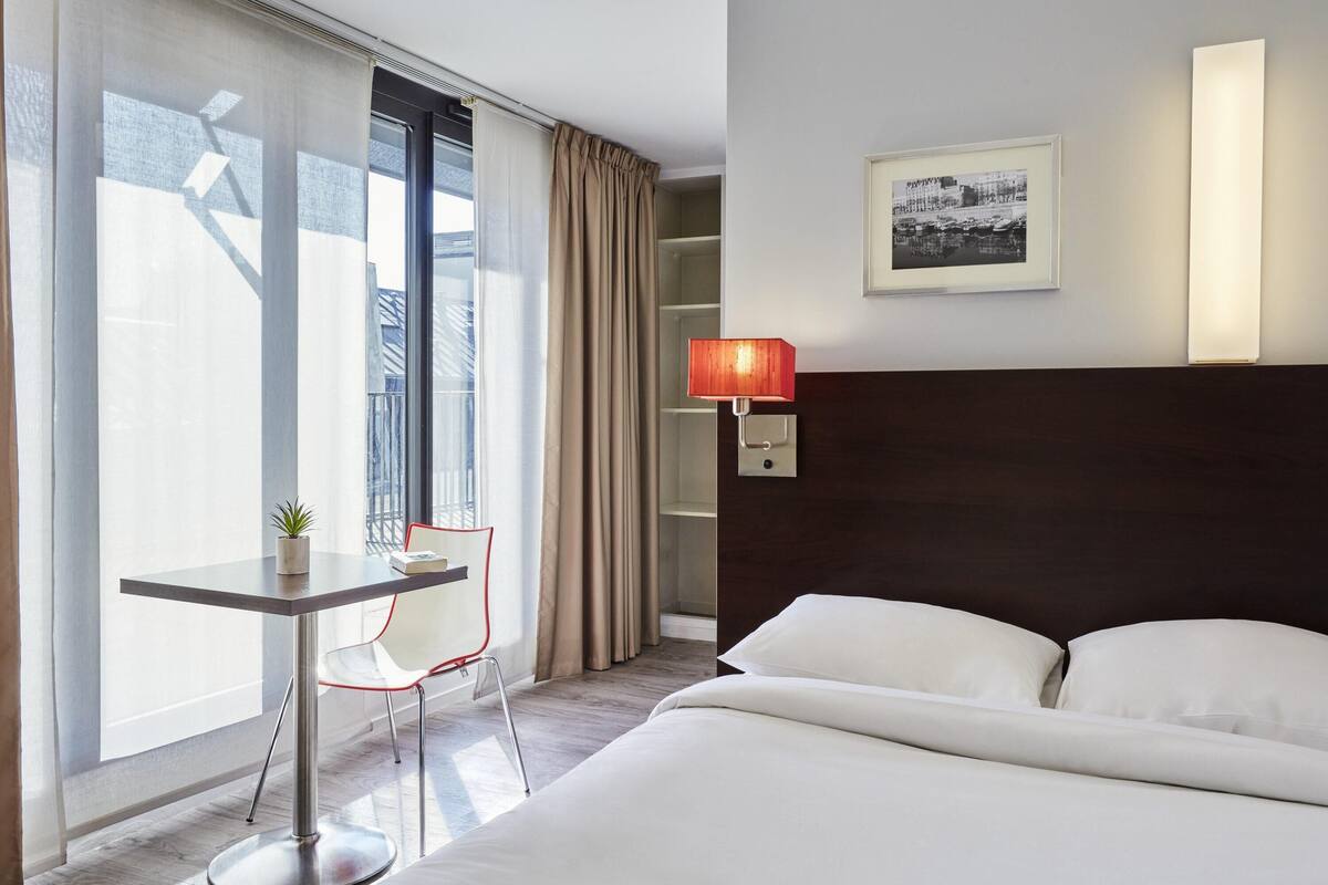Quarto de hotel com uma cama, uma escrivaninha com cadeira, uma janela com cortinas, uma luminária na parede e um quadro emoldurado.