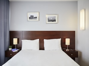 A hotel room with a large bed, two bedside tables, a dark wooden headboard, two framed pictures on the wall, and a nightstand with a lamp and a plant.