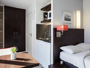 Una habitación de hotel compacta con kitchenette, una cama con ropa de cama blanca, una mesita de noche pequeña, una lámpara roja de pared y un cuadro enmarcado.