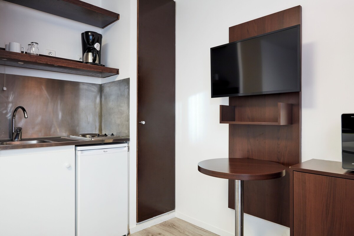 A compact kitchen with a dishwasher, a coffee maker, and a flat-screen TV mounted on a wooden wall.