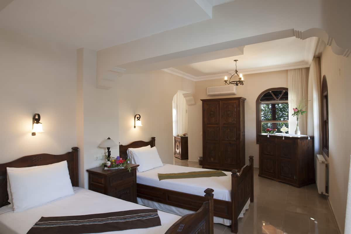 Habitación de hotel con dos camas, un armario de madera, un escritorio con lámpara y ventana con vista a follaje.