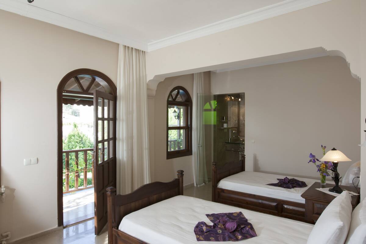 Habitación de hotel con dos camas, un balcón, baño con puerta de cristal y una mesita de noche con lámpara.