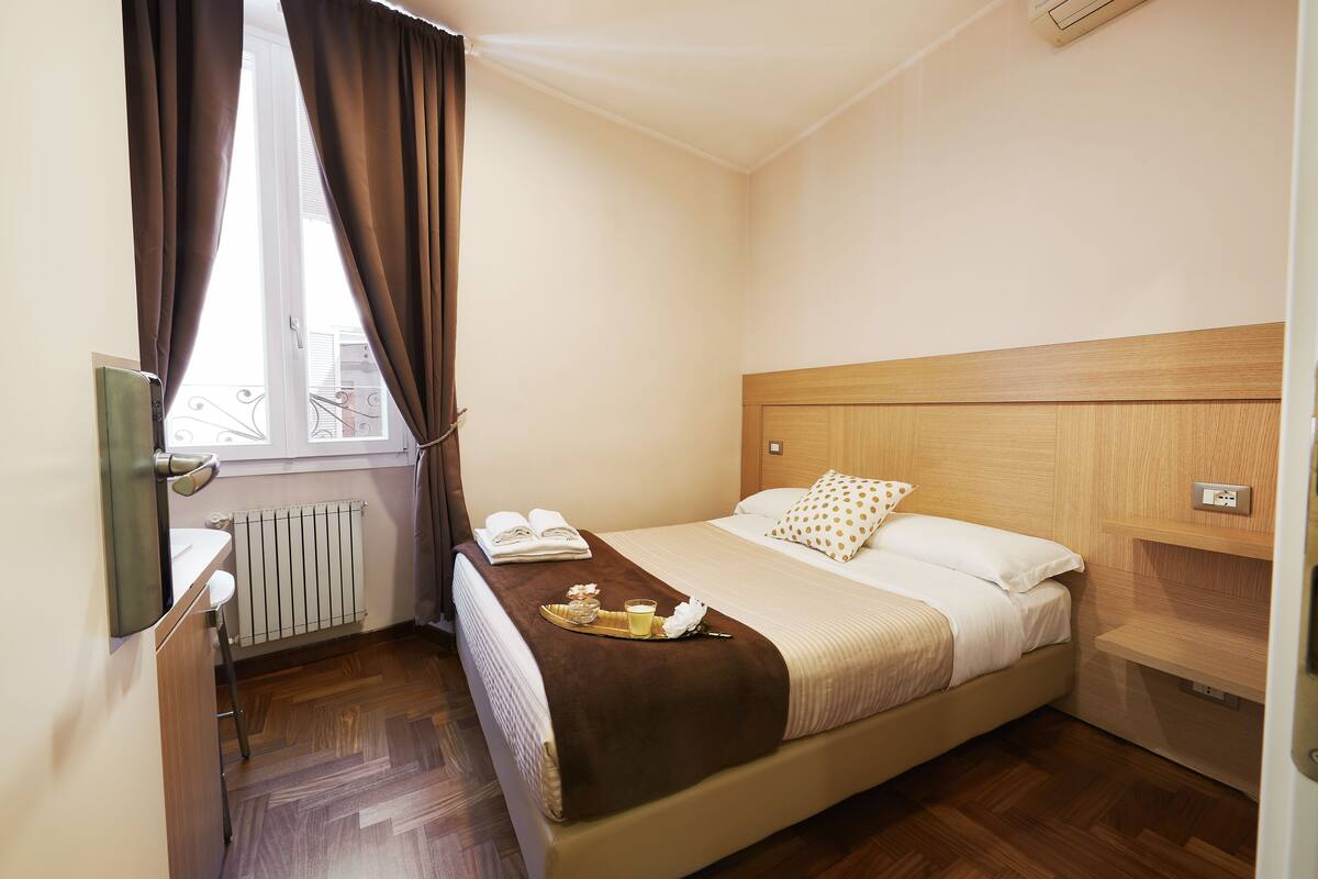 A hotel room with a bed, a bedside table, a radiator, and a window with curtains.