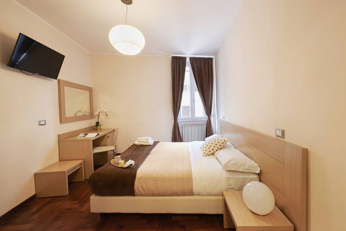 A neatly arranged hotel room with a bed, bedside table, a desk, a chair, a television mounted on the wall, and a window with curtains.