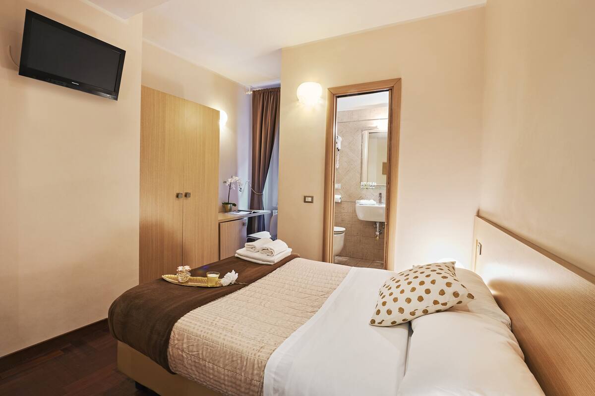 A hotel room with a bed, a TV mounted on the wall, a bathroom with a sink and mirror, and a door leading to another room.