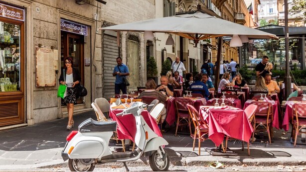 An outdoor cafe with tables and chairs, a scooter parked in front, and a building with a sign that reads "Pizza". - La Piccola Maison (Rome)