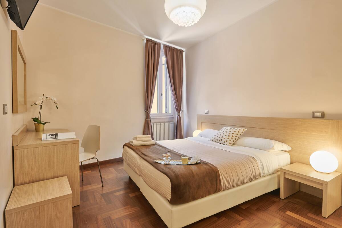 A modern hotel room with a wooden floor, a bed with a brown blanket, a bedside table with a lamp, a desk with a chair, and a window with curtains.