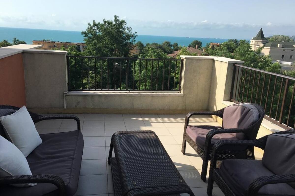 A balcony with wicker furniture, a view of the sea, and surrounding buildings.