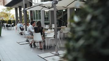 Un café en plein air avec des tables et des chaises blanches, de grands parasols et un bâtiment moderne en arrière-plan.
