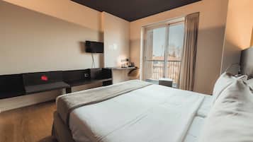 Ein Hotelzimmer mit einem großen Bett, einem an der Wand montierten Fernseher, einem kleinen Tisch mit einer Lampe und einem Fenster mit Vorhängen.
