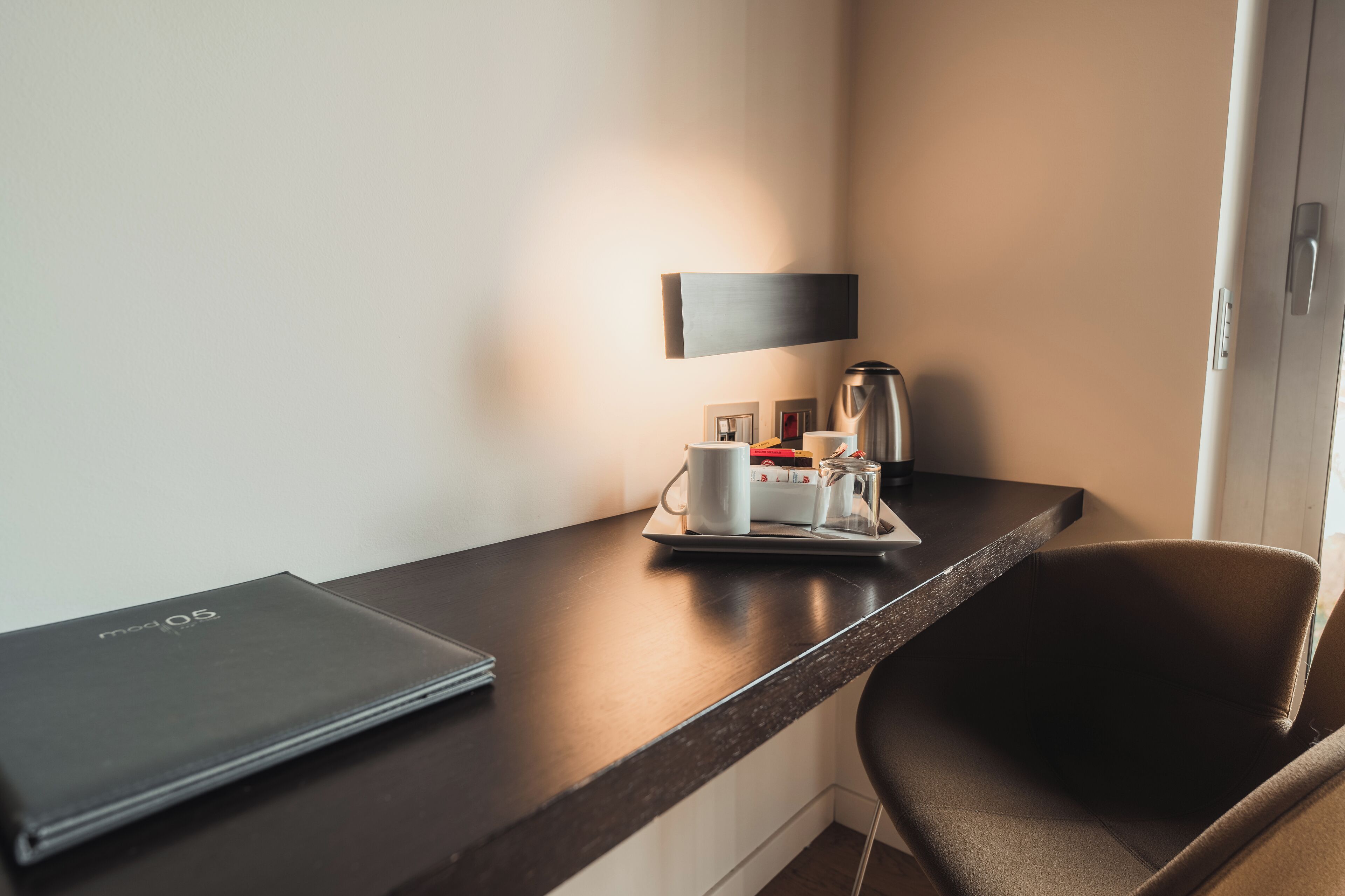Ein Hotelzimmer mit dunklem Holztisch, einem Laptop, einer Ablage mit Kaffeekocher, Tassen und einer kleinen Tischlampe.