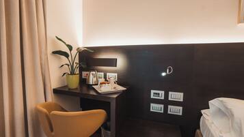 Quarto de hotel com escrivaninha, cadeira, cama, planta em vaso e mesa de cabeceira com abajur e uma xícara.