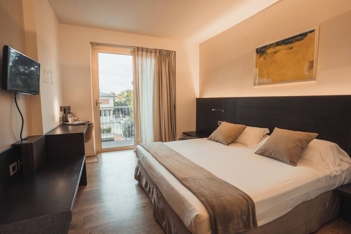 A modern hotel room with a large bed, a flat-screen TV, a desk, and a balcony with curtains.