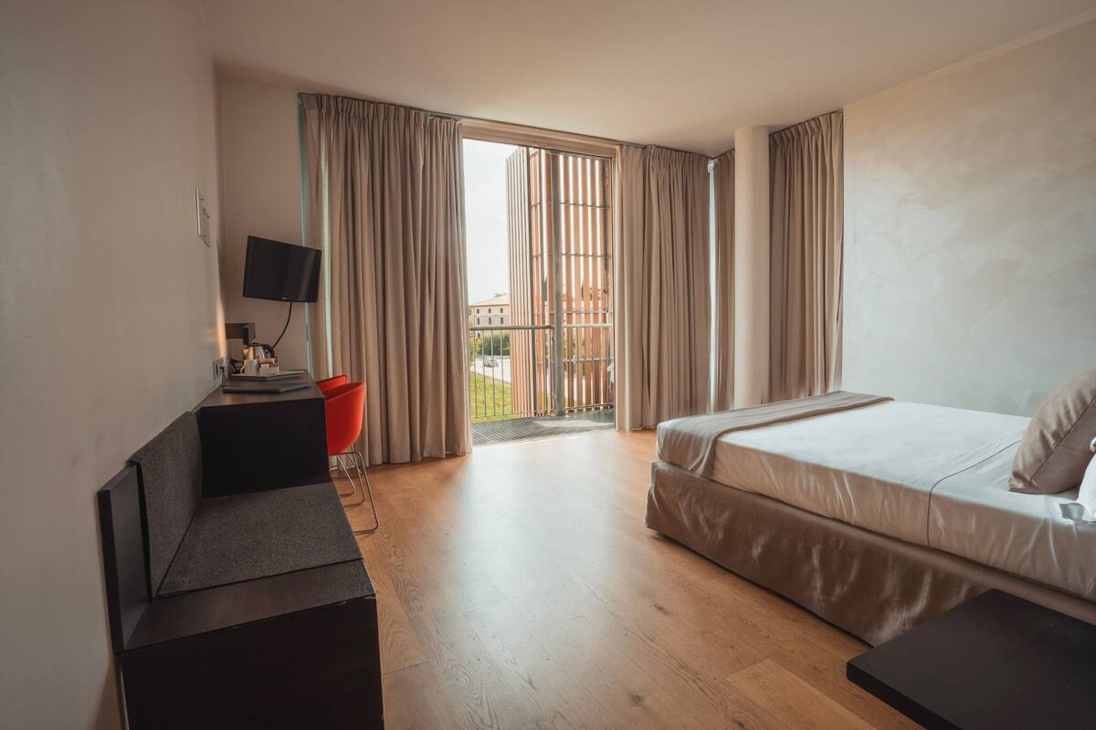 A modern hotel room with a bed, a desk, a television, and a view of the outside.
