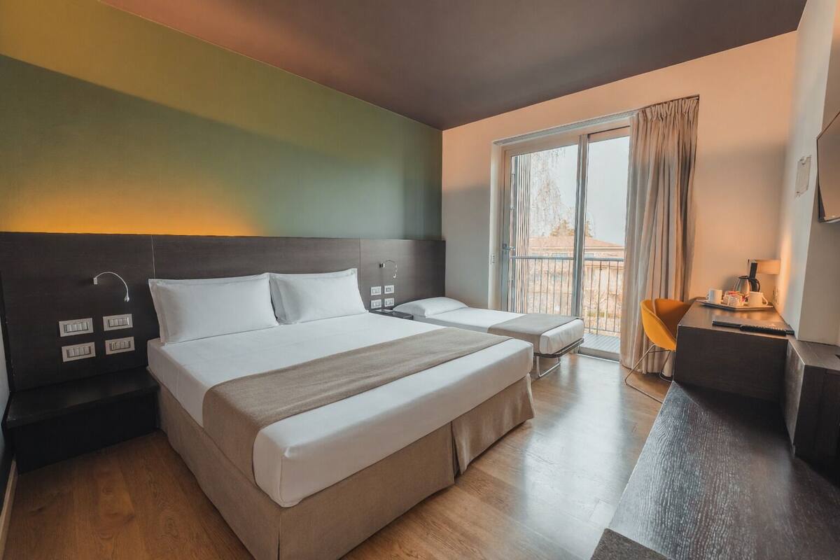 A hotel room with a large bed, a smaller bed, a desk with a chair, and a window with curtains.