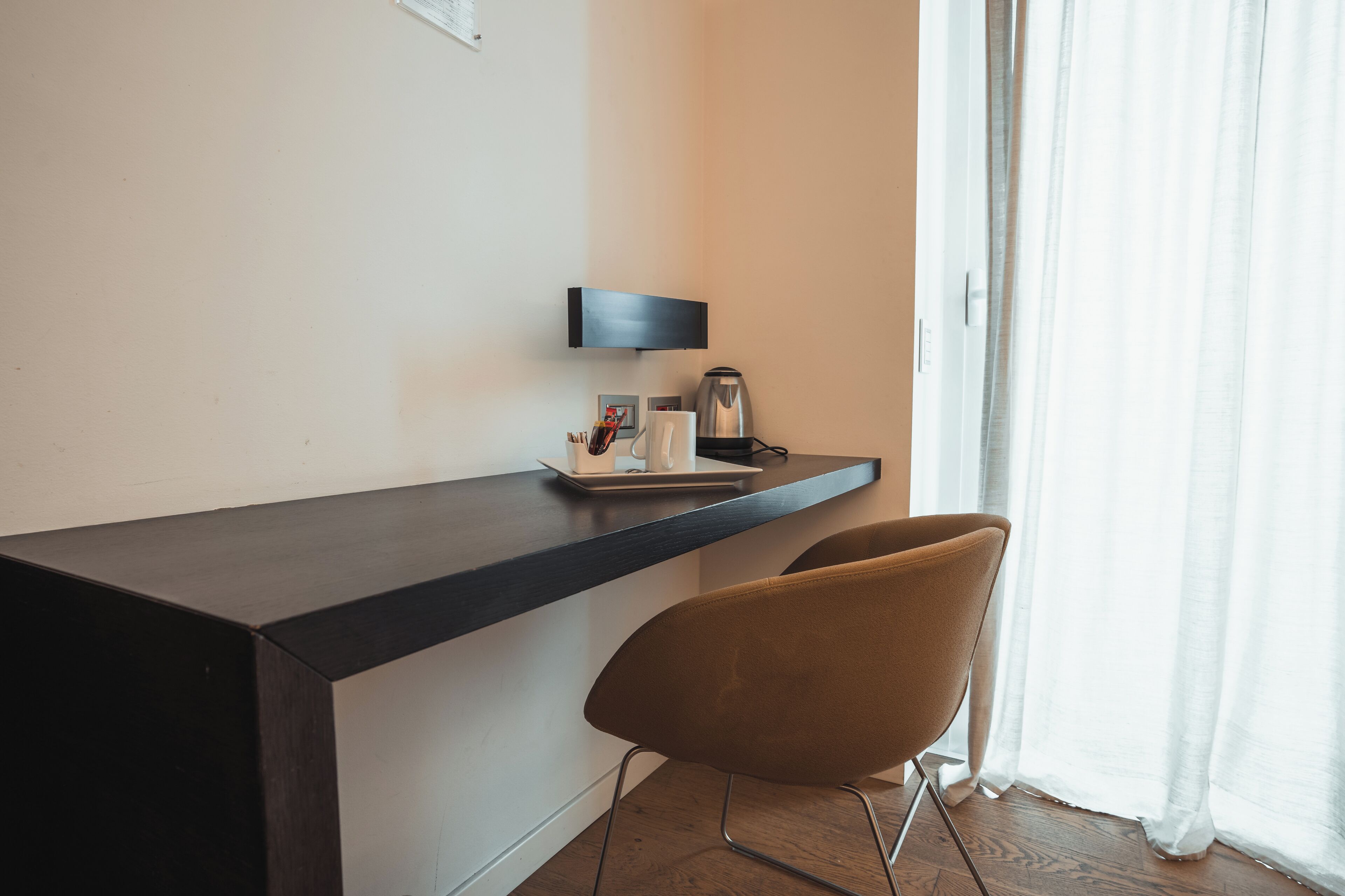 Une chambre moderne avec un bureau en bois foncé, une chaise marron, une lampe fixée au mur et une fenêtre ornée de rideaux légers.