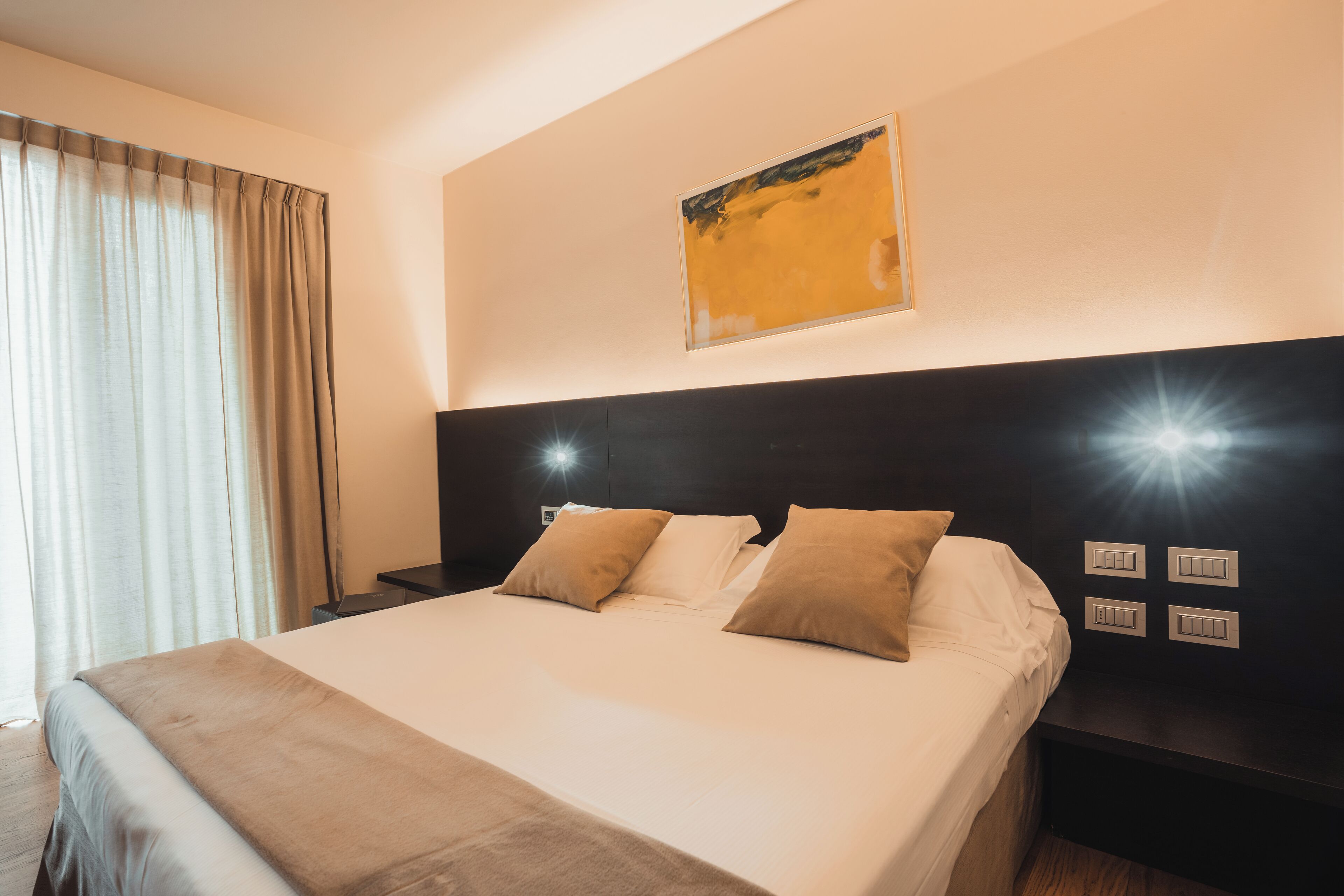 Habitación de hotel con una cama grande, cabecera con interruptores de luz, mesita de noche y ventana con cortinas.