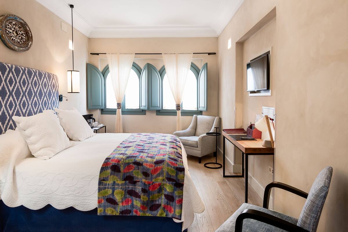 A hotel room with a bed, a desk, a chair, a lamp, and a vase with flowers.