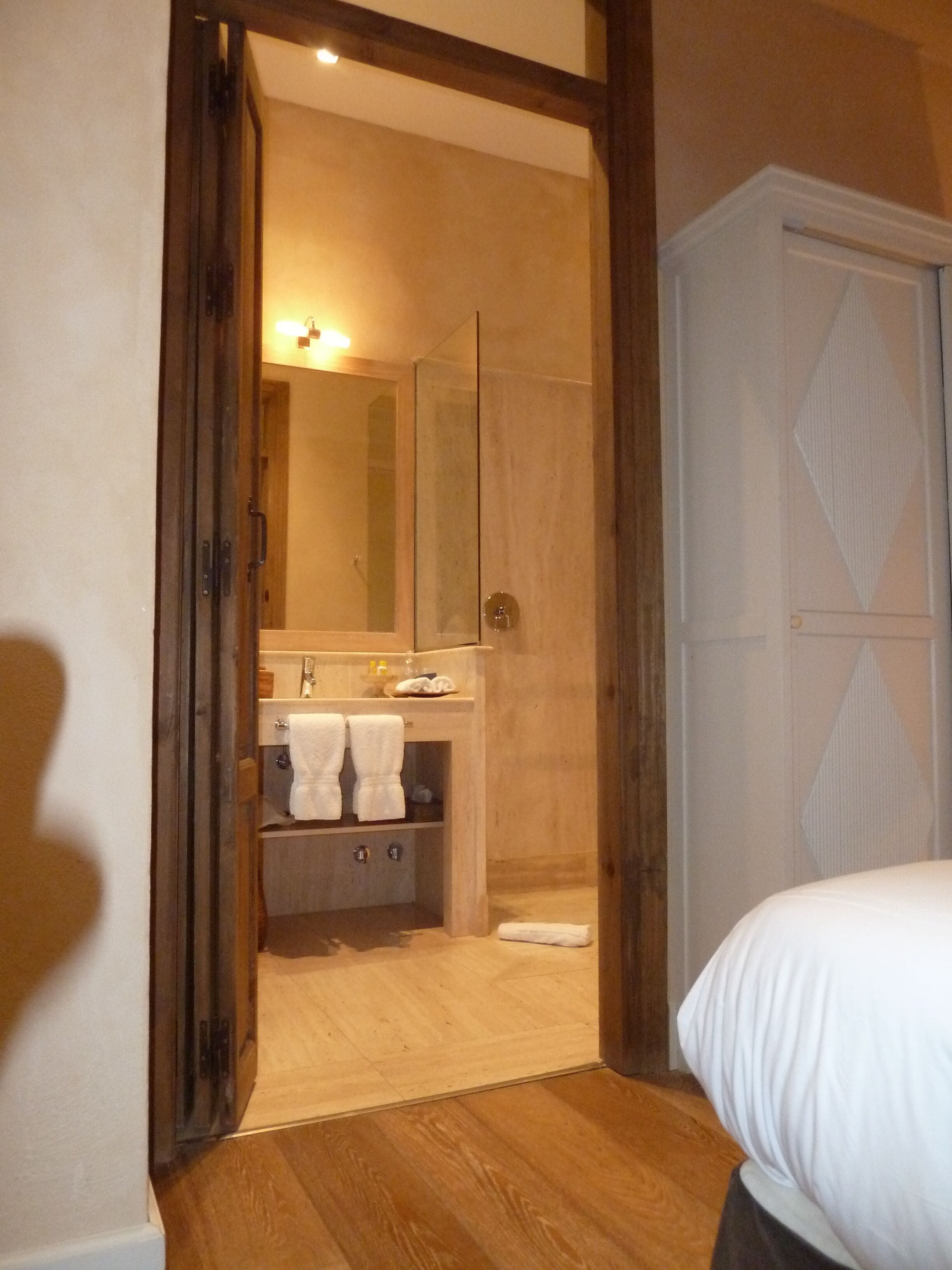 Un dormitorio con suelo de madera, una cama, un armario y un baño con espejo, lavabo y toallas.