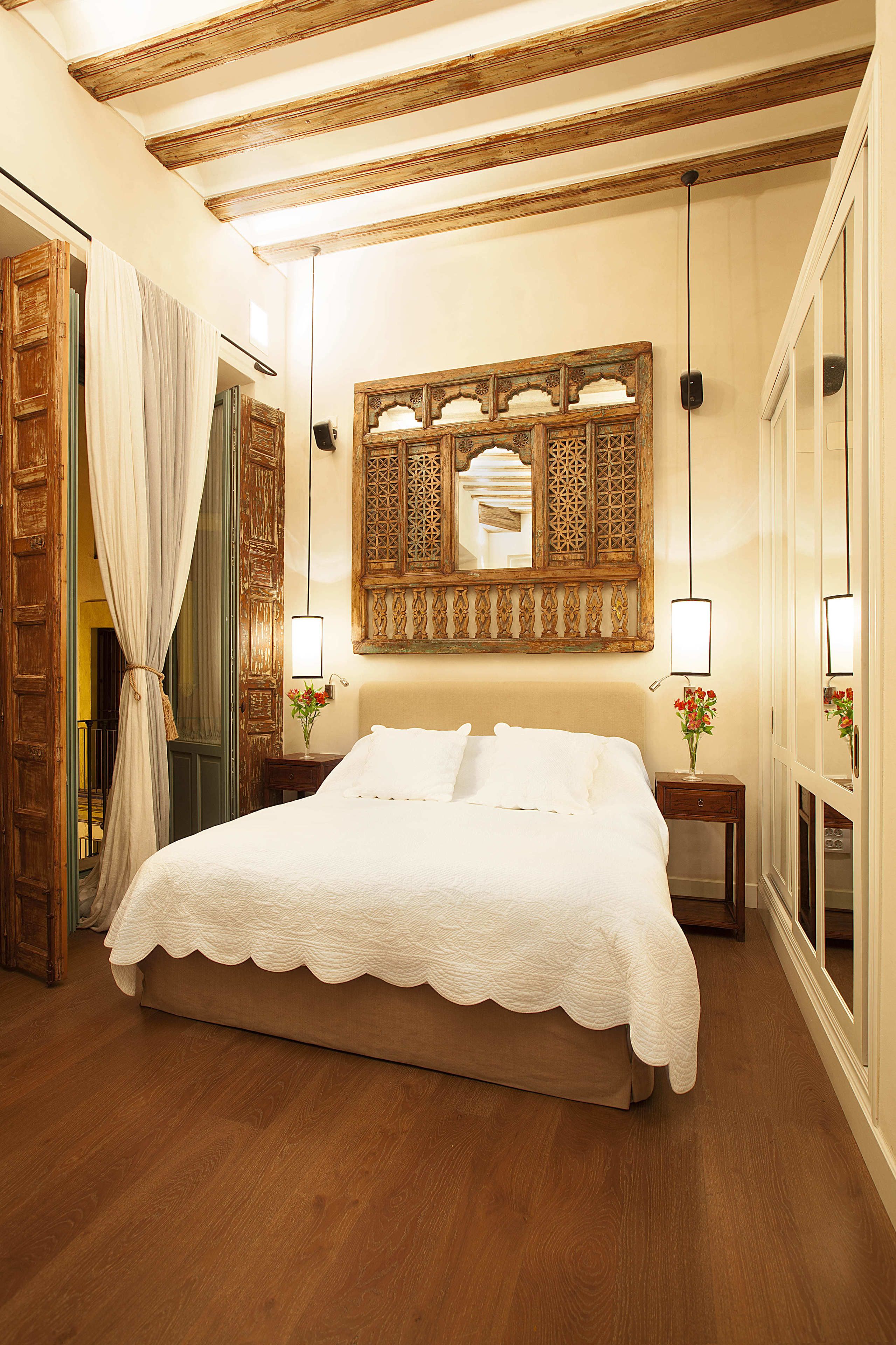 Quarto com cama grande, vigas de madeira no teto, espelho decorativo e duas luminárias de cabeceira.