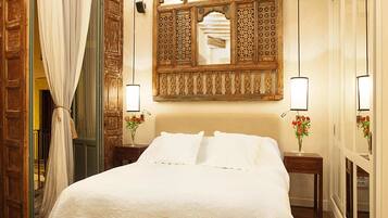 Quarto com cama grande, vigas de madeira no teto, espelho decorativo e duas luminárias de cabeceira.