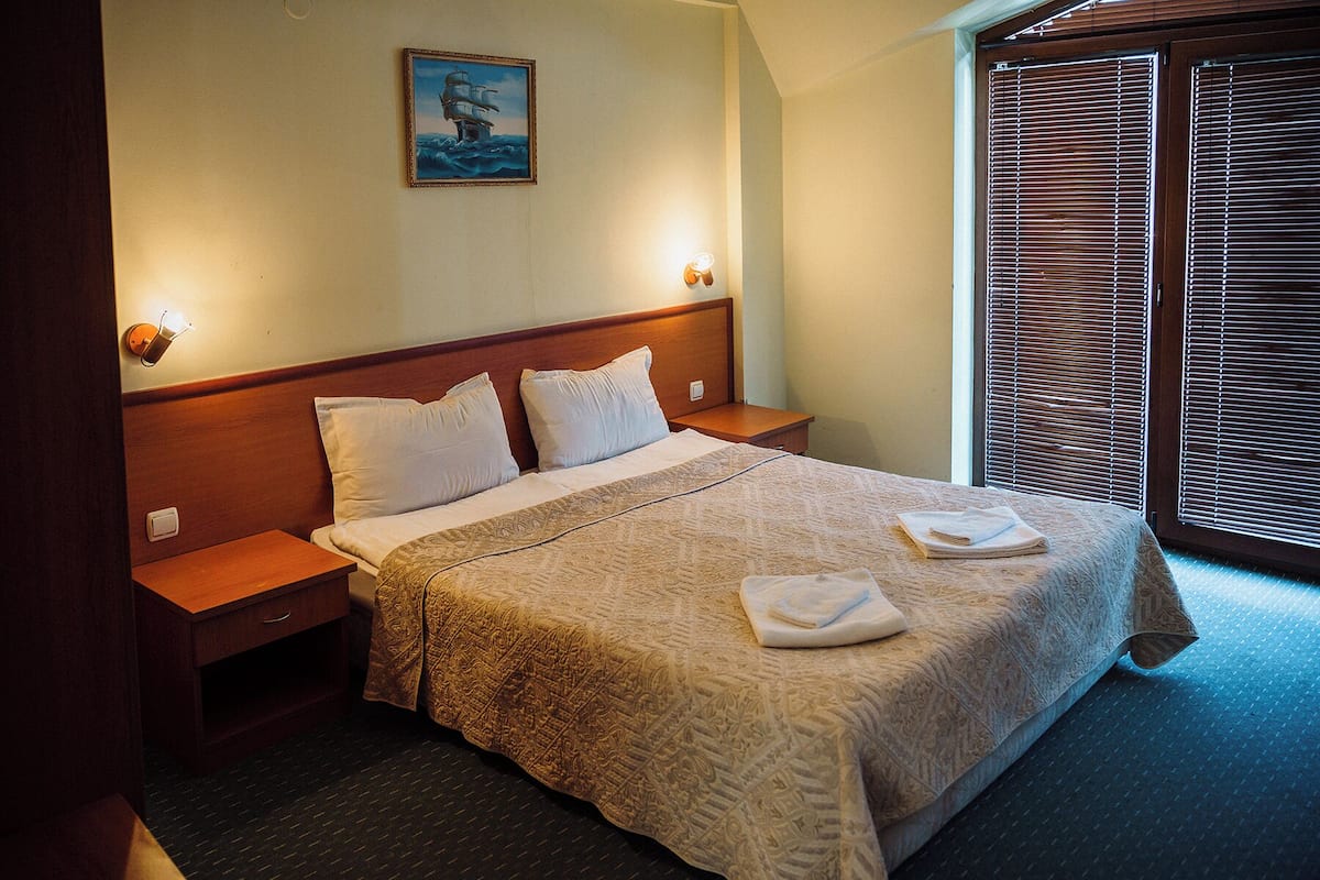 A neatly made bed with a patterned bedspread, two pillows, and two folded towels on the bed. There is a nightstand with a lamp on it, and a painting of a ship on the wall.