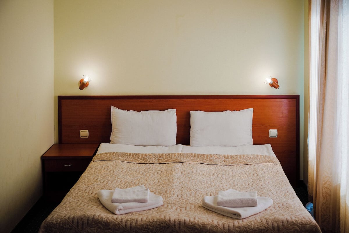 A hotel room with a wooden headboard, two white pillows, a patterned bedspread, and two folded towels on the bed.