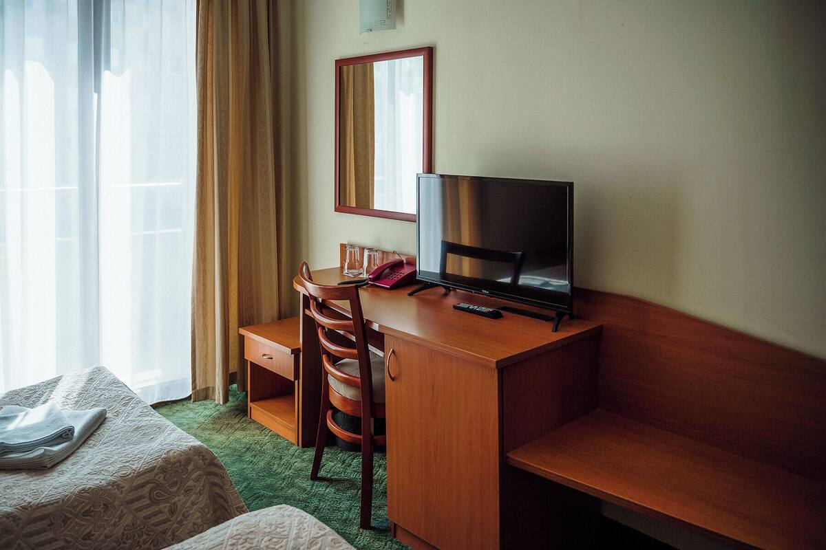 A hotel room with a wooden desk, a television, a chair, a bed, and a window with curtains.