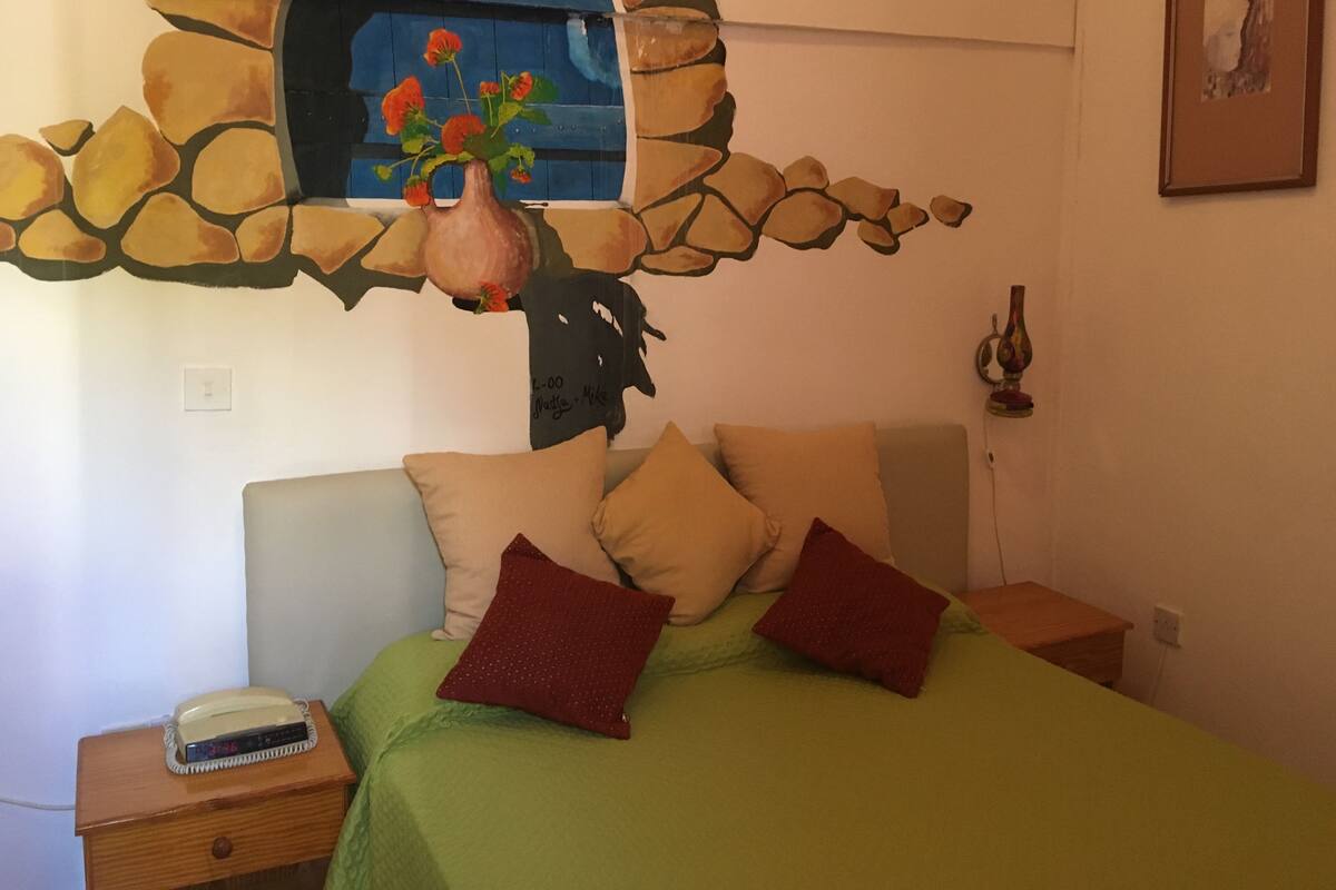 A bedroom with a bed, pillows, a nightstand, a lamp, and a painting of flowers on the wall.