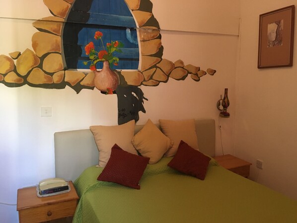 Une chambre à coucher avec un lit, des oreillers, une table de chevet, une lampe et un tableau représentant des fleurs accroché au mur.