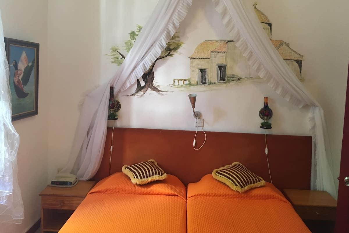 A bedroom with a bed, two pillows, a nightstand, a wall clock, a framed picture, and a decorative wall hanging.
