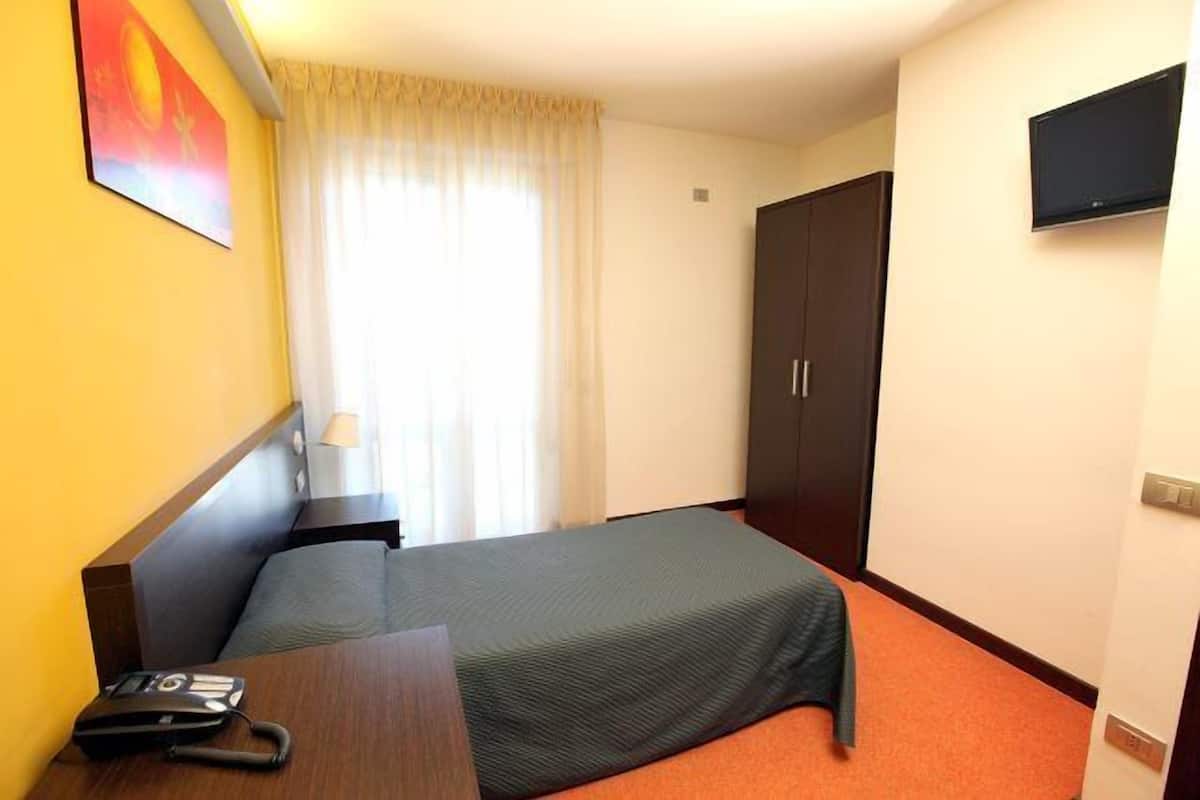 A hotel room with a bed, a desk with a telephone, a television mounted on the wall, and a wardrobe.