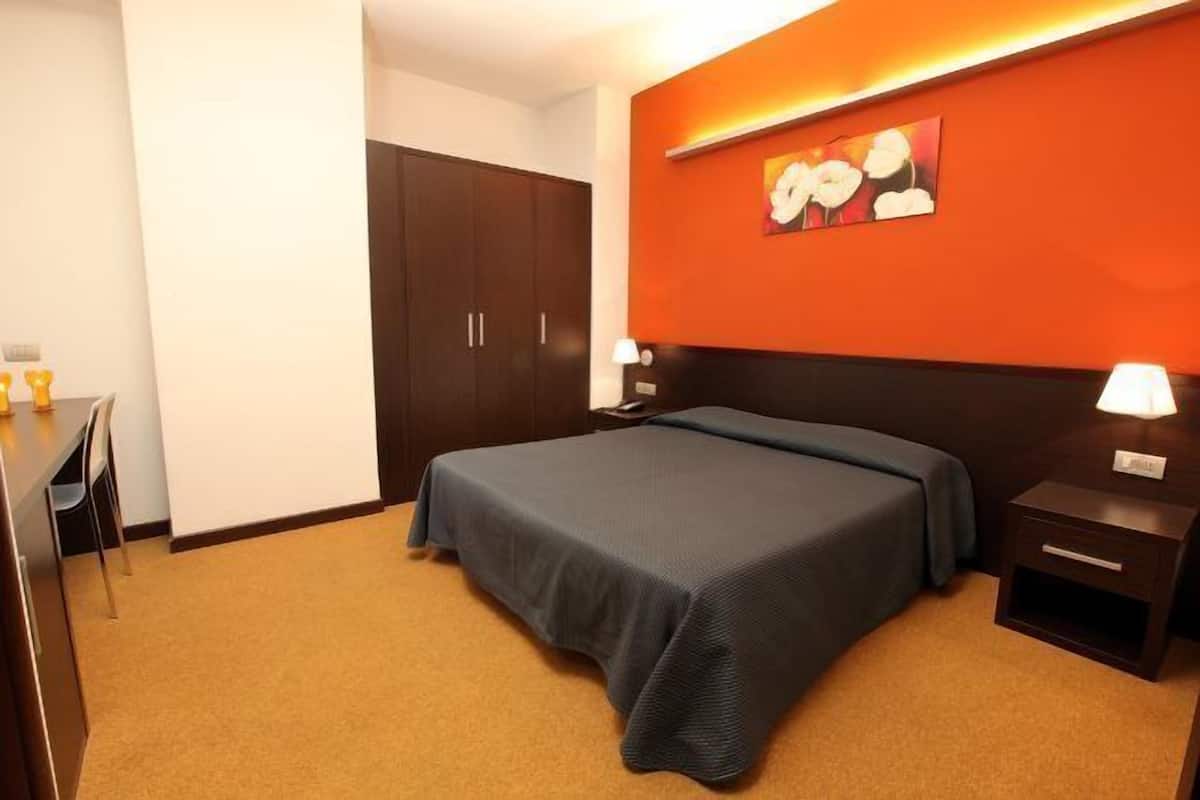 A hotel room with a bed, a desk, a chair, a wardrobe, and a nightstand with a lamp.