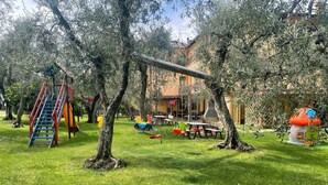 A playground with a slide and climbing structure, surrounded by trees and a building with outdoor seating. - Casa Chincarini (Malcesine)