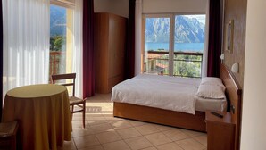 A bedroom with a bed, a chair, a table, and a view of mountains and a lake. - Casa Chincarini (Malcesine)
