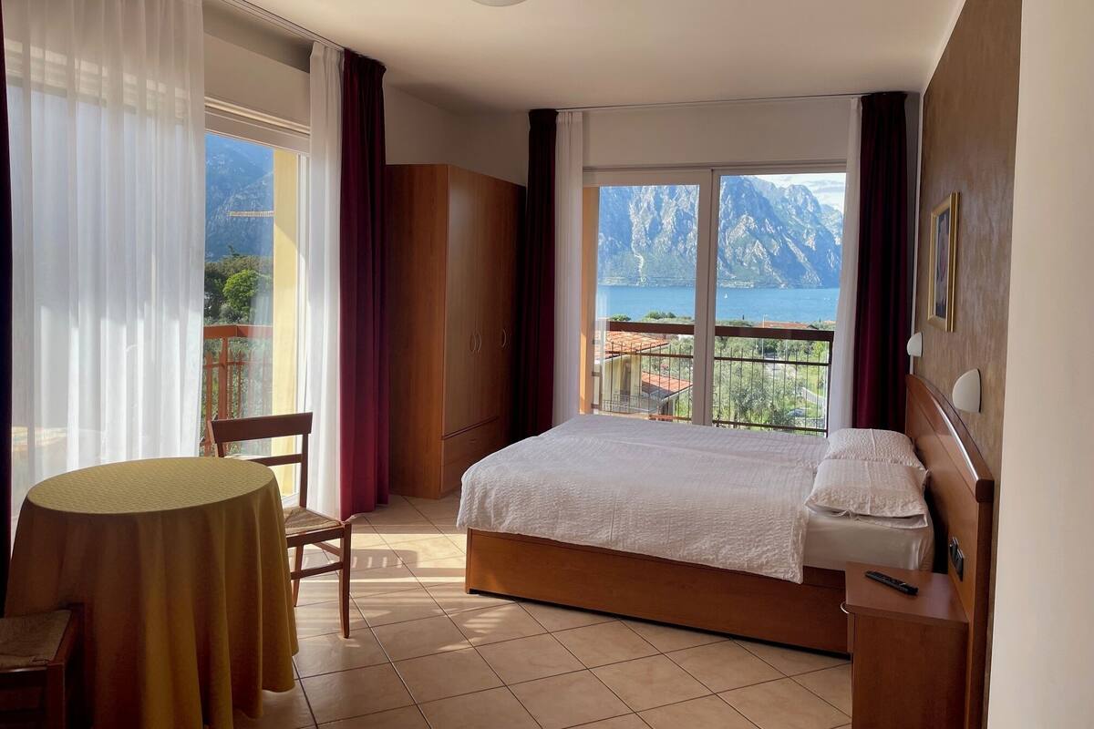 A bedroom with a bed, a chair, a table, and a view of mountains and a lake.