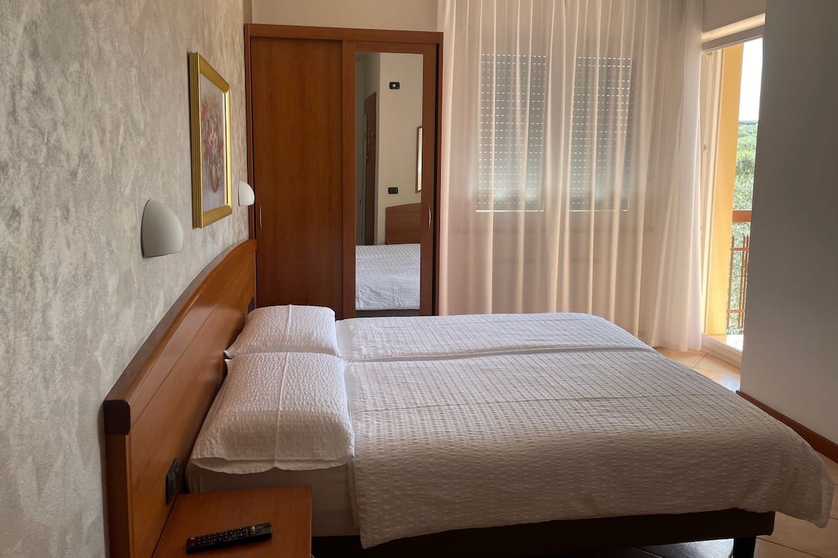 A hotel room with a wooden bed, a bedside table with a remote control, a mirror, and a view of the outside through sheer curtains.