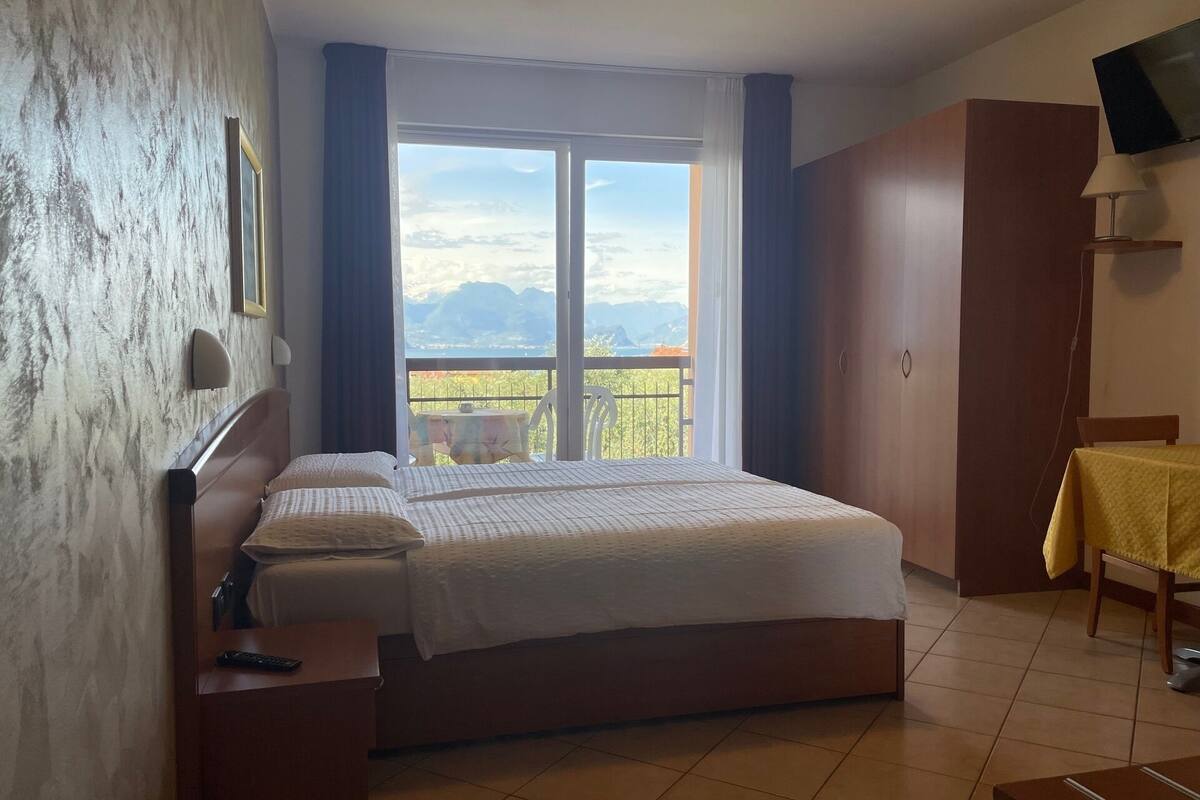 A hotel room with a bed, a desk, a chair, a TV, and a balcony with a view of mountains.