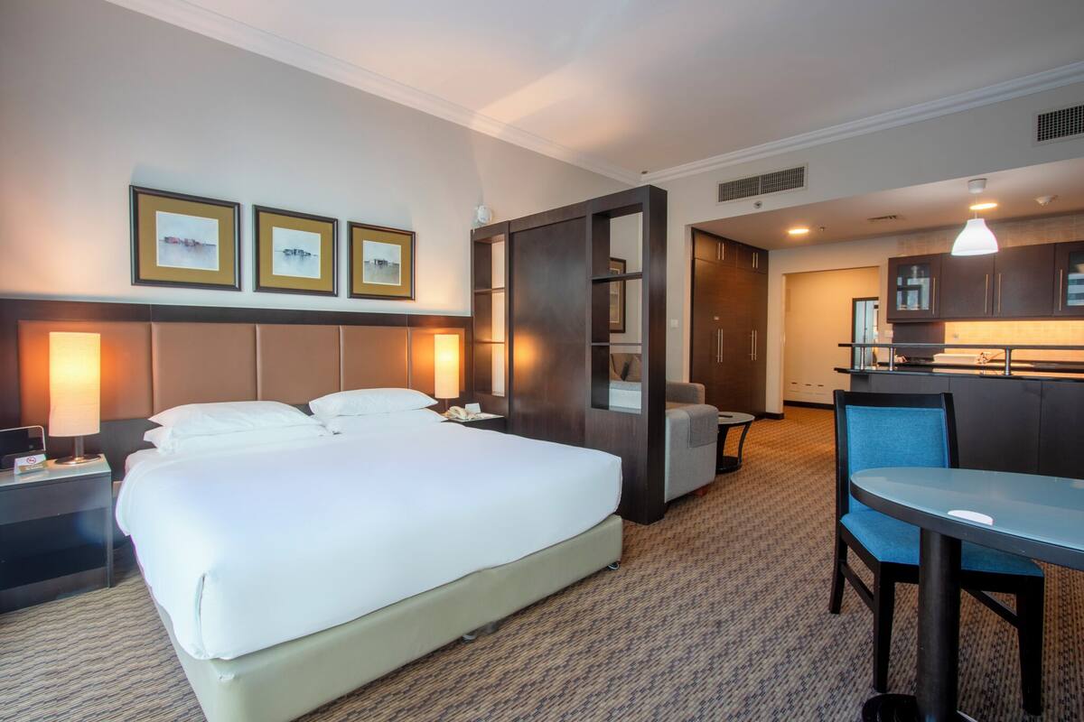 Habitación de hotel con una cama grande, lámparas de noche, un escritorio, una silla y una kitchenette.