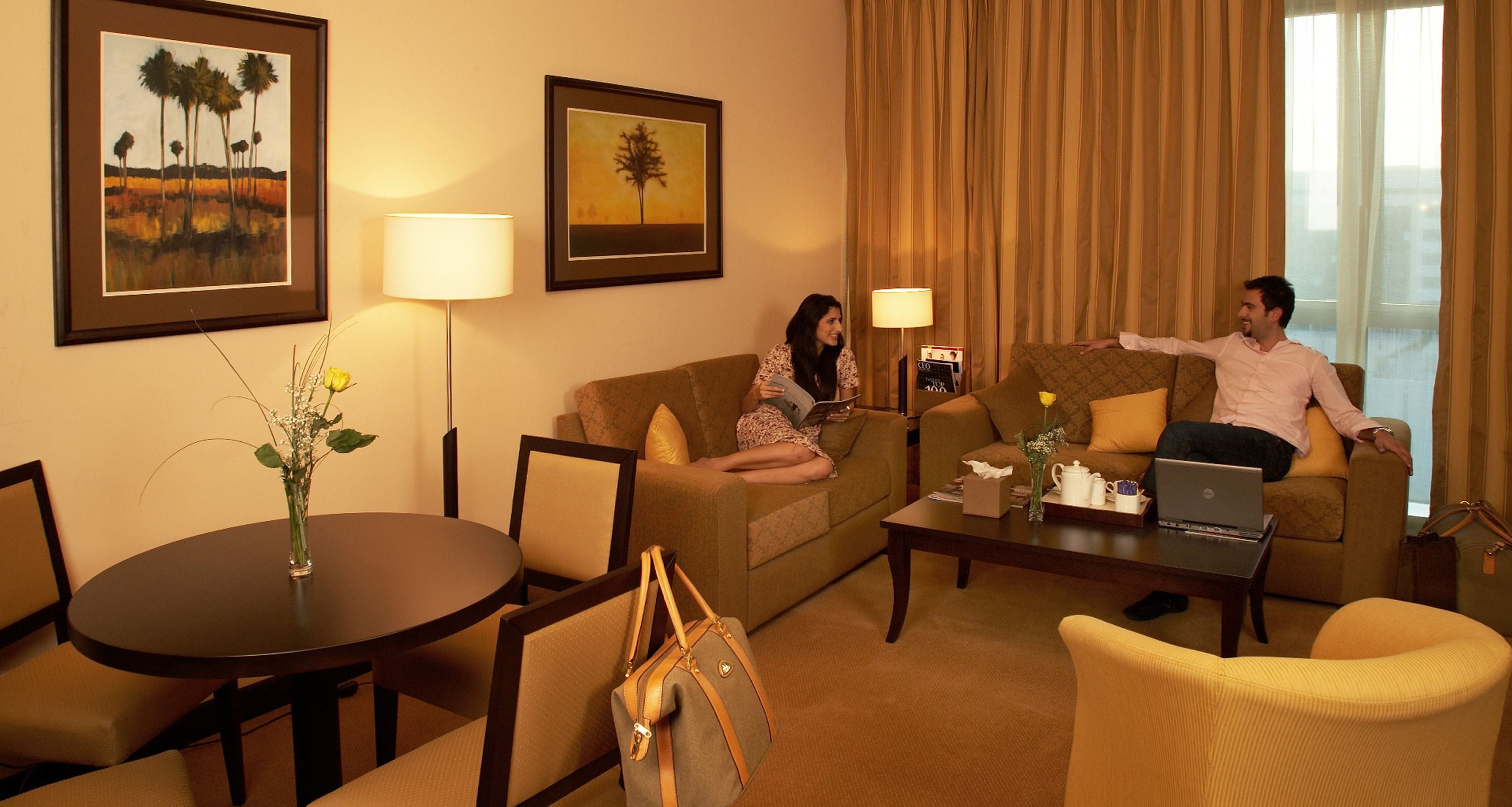 A hotel room with a sofa, armchairs, a coffee table, a dining table, and a vase with flowers.
