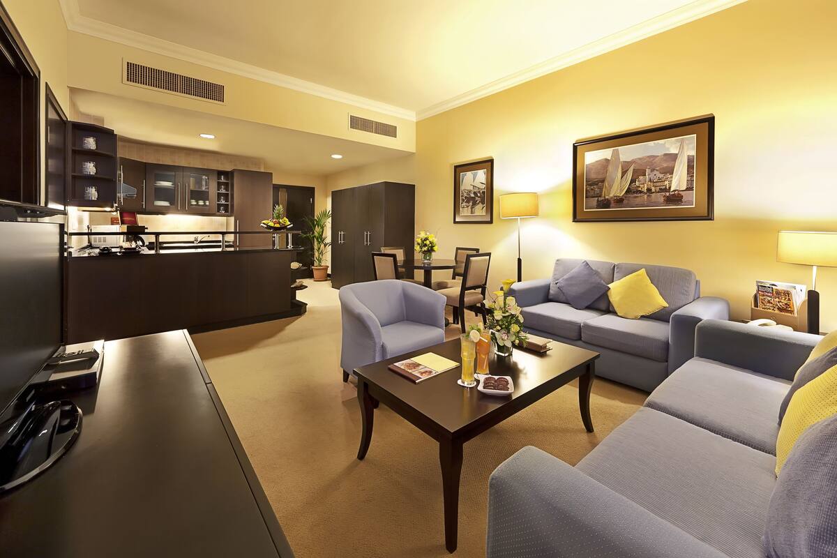 Una habitación de hotel moderna con sala de estar, una mesa de comedor, un conjunto de sofás, una zona de cocina y un televisor.