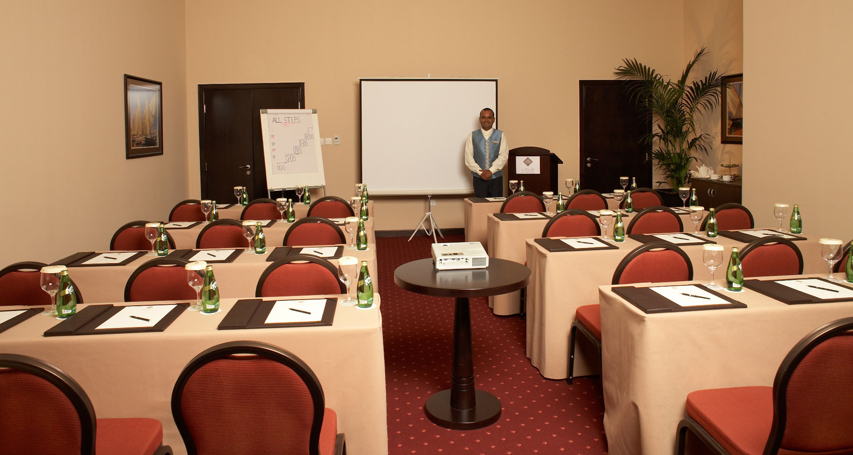 Une salle de conférence avec des tables disposées pour une réunion, un tableau blanc et un présentateur à un pupitre.