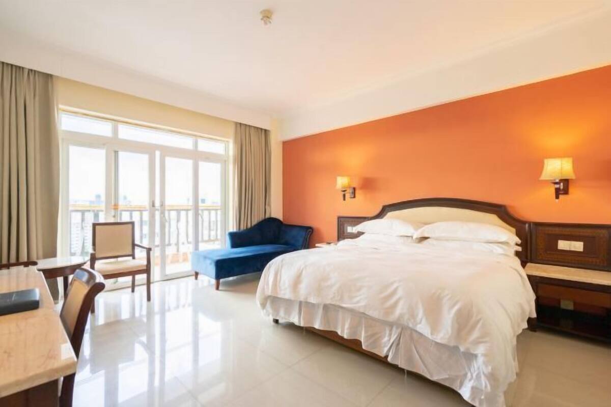 Quarto de hotel com uma cama grande, uma escrivaninha, uma cadeira, uma varanda e duas luminárias fixadas na parede.