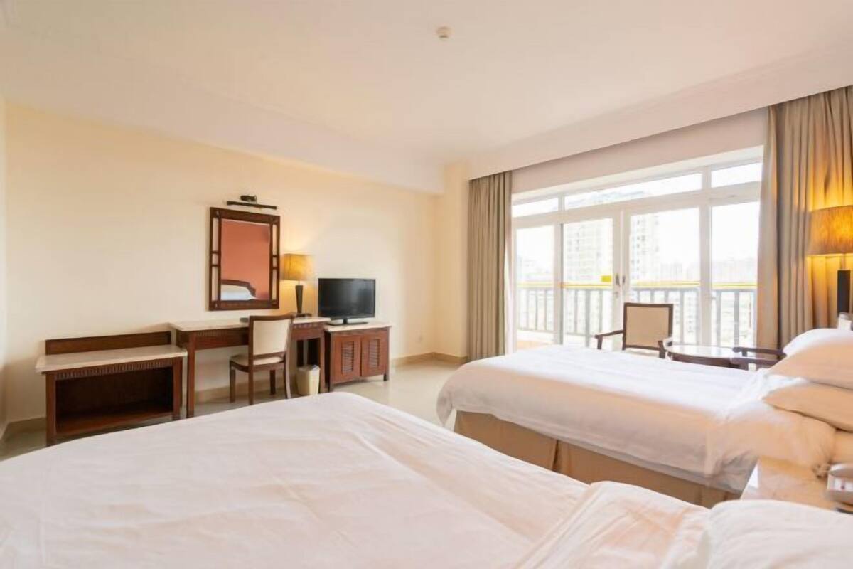 Quarto de hotel com duas camas, uma escrivaninha com cadeira, televisão e varanda com vista.